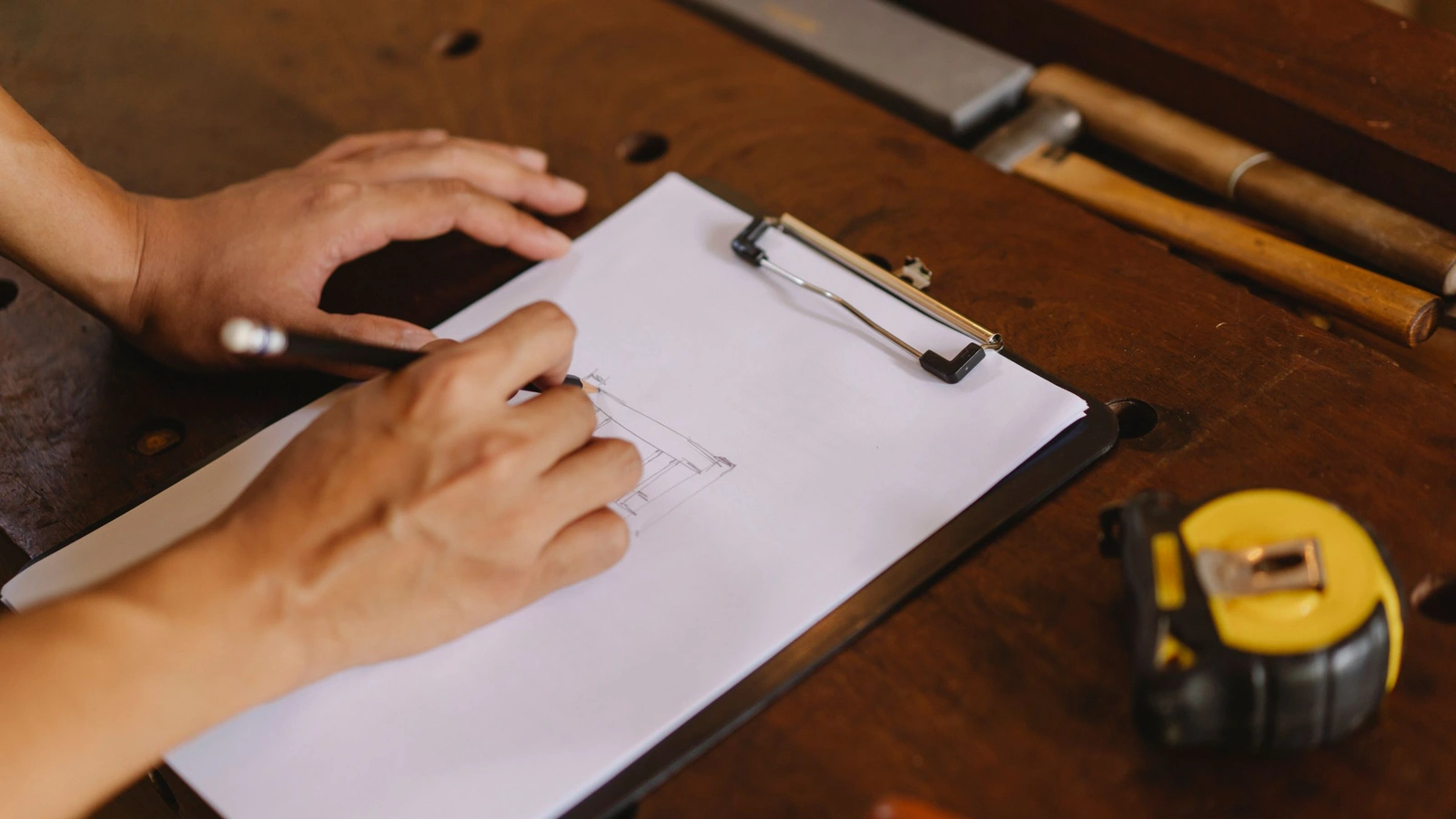 Mains traçant un croquis au crayon sur une planchette à pince, à côté d'un mètre ruban sur un établi