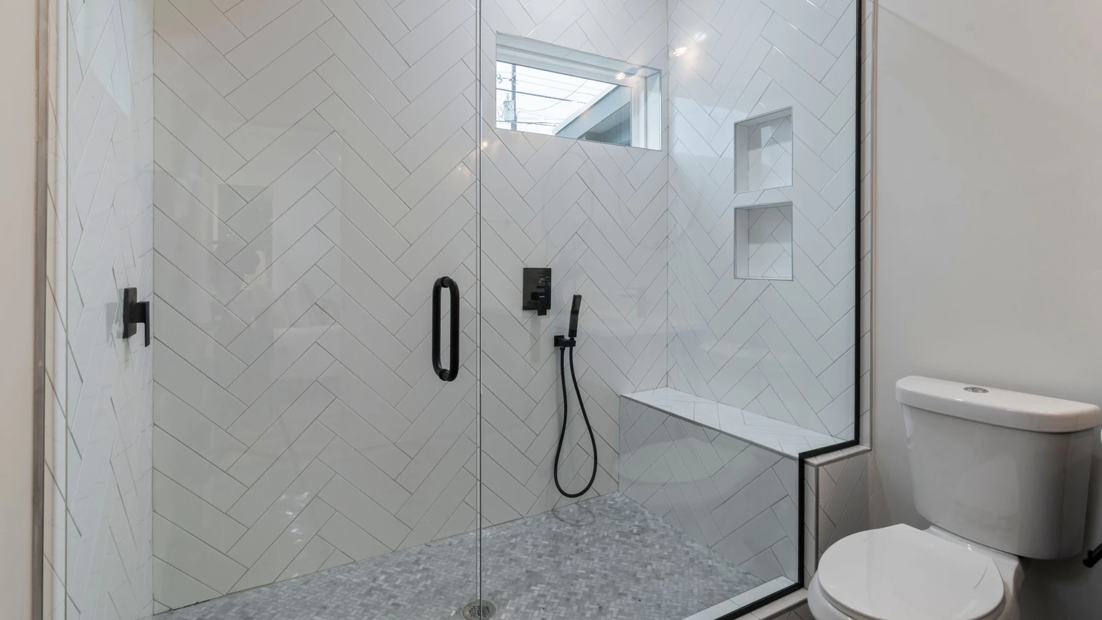 Douche vitrée au carrelage blanc posé en chevrons, avec un banc maçonné et une robinetterie noire
